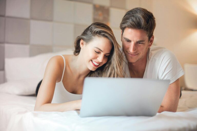 Uśmiechnięta para leży na łóżku obok siebie i razem patrzy w ekran laptopa
