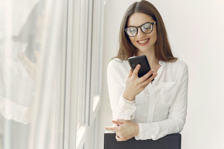 Uśmiechnięta kobieta w okularach i białej koszuli patrzy na telefon, stojąc przy dużym oknie