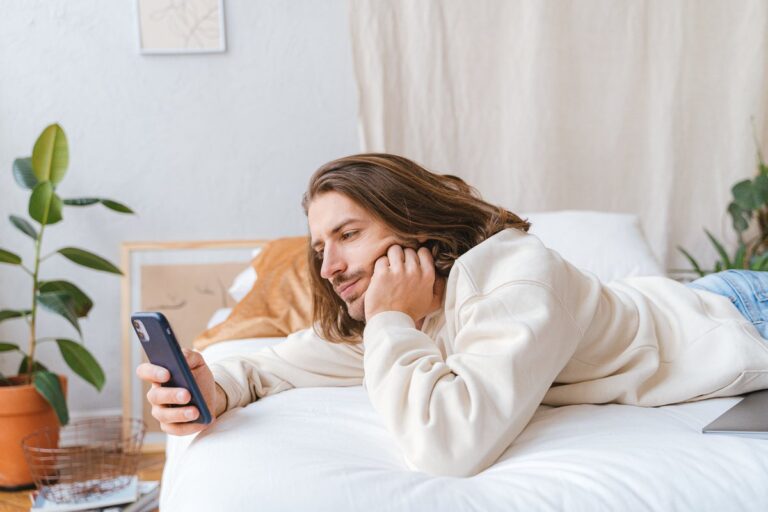 Długowłosy mężczyzna leży na łóżku, podpiera głowę ręką i patrzy w telefon, obok roślina doniczkowa